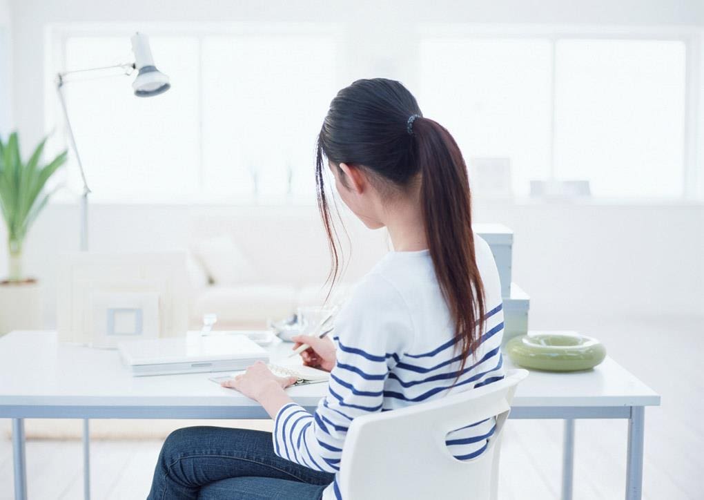 A woman in a striped sweater writing at a bright desk.